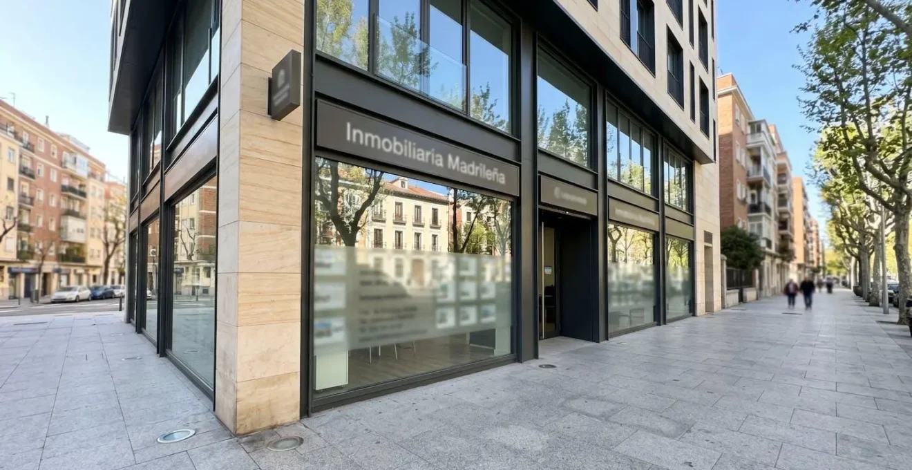 Fachada de una oficina inmobiliaria contemporánea en calle peatonal luminosa de barrio residencial, arquitectura europea moderna con escaparate de cristal, personas difuminadas al fondo