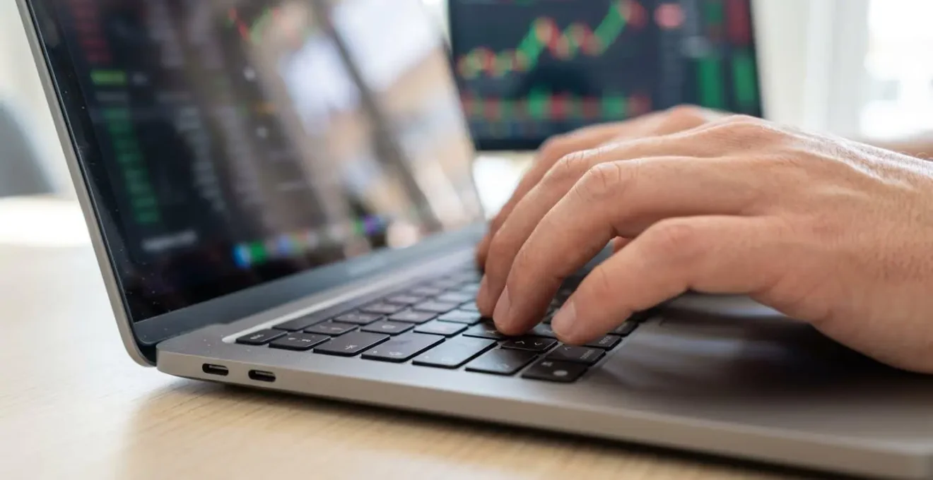 Primer plano de manos tecleando en teclado de portátil moderno con pantalla mostrando gráficos borrosos al fondo